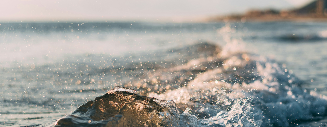 Close-up of ocean waves splashing toward the camera, with sparkling water droplets in soft light and a blurred shoreline in the distance.